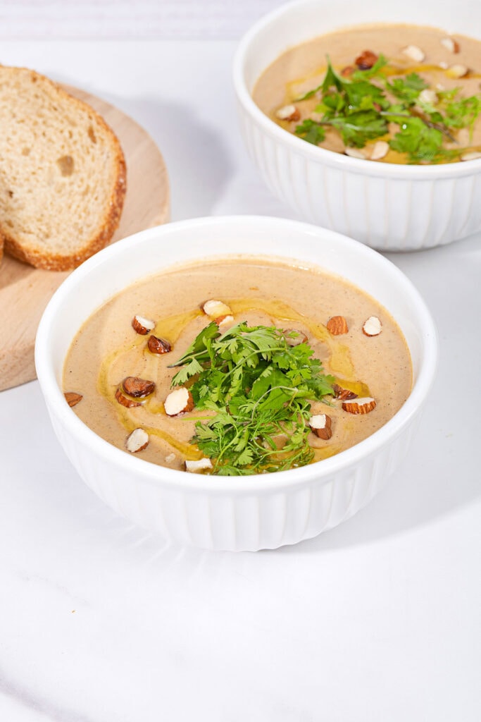 On a marbled table sits two white bowls of creamy vegan cauliflower soup, topped with olive oil, sliced almonds and cilantro. Next to them is a board with some slices of bread | Hurry The Food Up