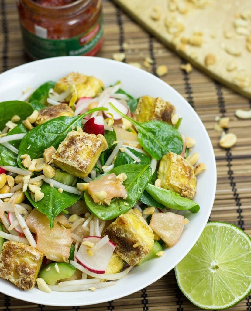 Asian tofu salad is served in a white bowl; a jar of sriracha, some crushed peanuts and a half lime are around it on the table | Hurry The Food Up