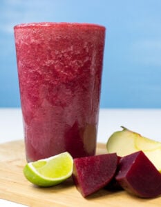 The glass with beetroot smoothie is on the wooden cut board. Near it, there are some pieces of beetroot, lime and apple | Hurry The Food Up