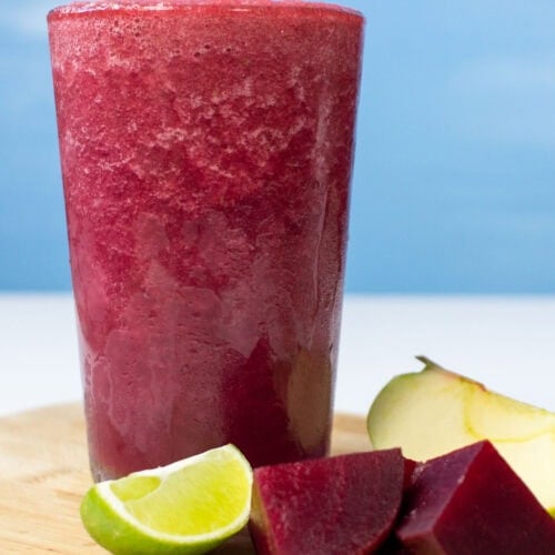 The glass with beetroot smoothie is on the wooden cut board. Near it, there are some pieces of beetroot, lime and apple | Hurry The Food Up