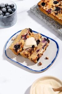 A few slices of baked oats topped with cream cheese frosting are on the plate surrounded with a marble plate with lemon berry baked oats, a glass of blueberries and a small bowl of cream cheese frosting | Hurry The Food Up