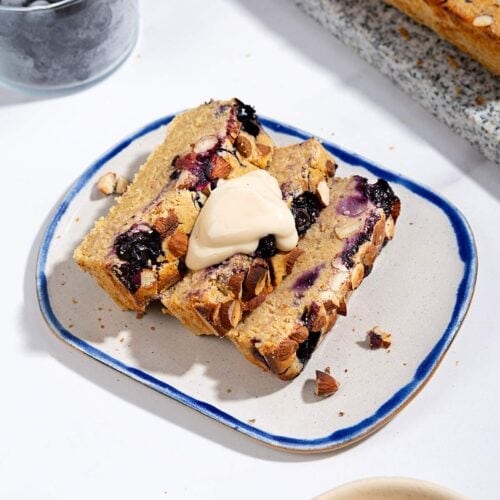 A few slices of baked oats topped with cream cheese frosting are on the plate surrounded with a marble plate with lemon berry baked oats, a glass of blueberries and a small bowl of cream cheese frosting | Hurry The Food Up