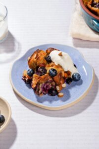 A portion of dessert served on a blue plate with a dollop of greek yogurt and a tray of blueberries. | Hurry The Food Up