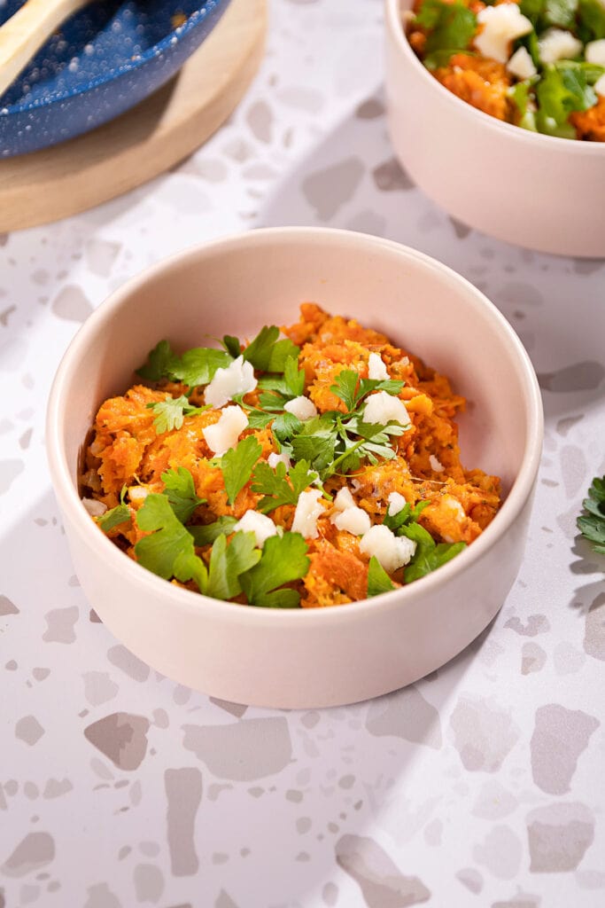 Two ceramic bowls sit on a grey table with the served cheesy sweet potato mash in. To the right there is some parsley, to the left in the background is a blue saucepan on a chopping board with a wooden spoon | Hurry The Food Up