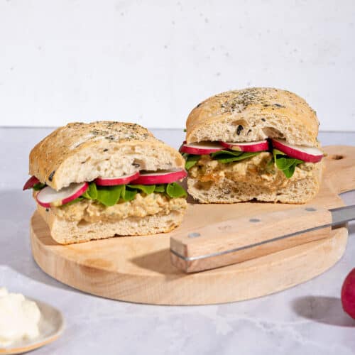 Two chickpea salad sandwiches with knife are on a wooden board that is on a marble surface | Hurry The Food Up