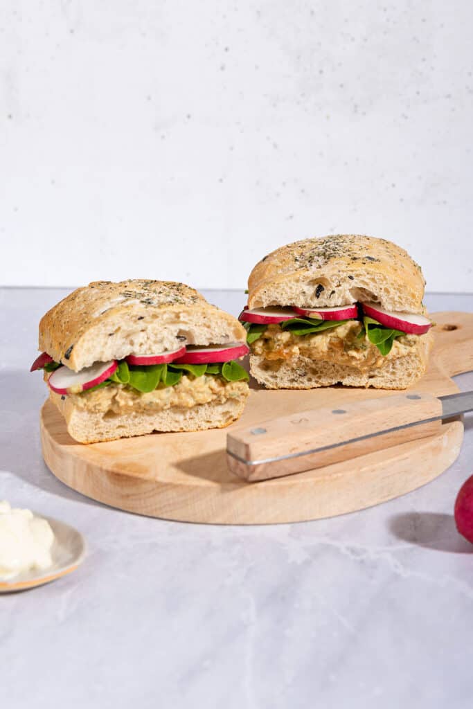 Two chickpea salad sandwiches with knife are on a wooden board that is on a marble surface | Hurry The Food Up