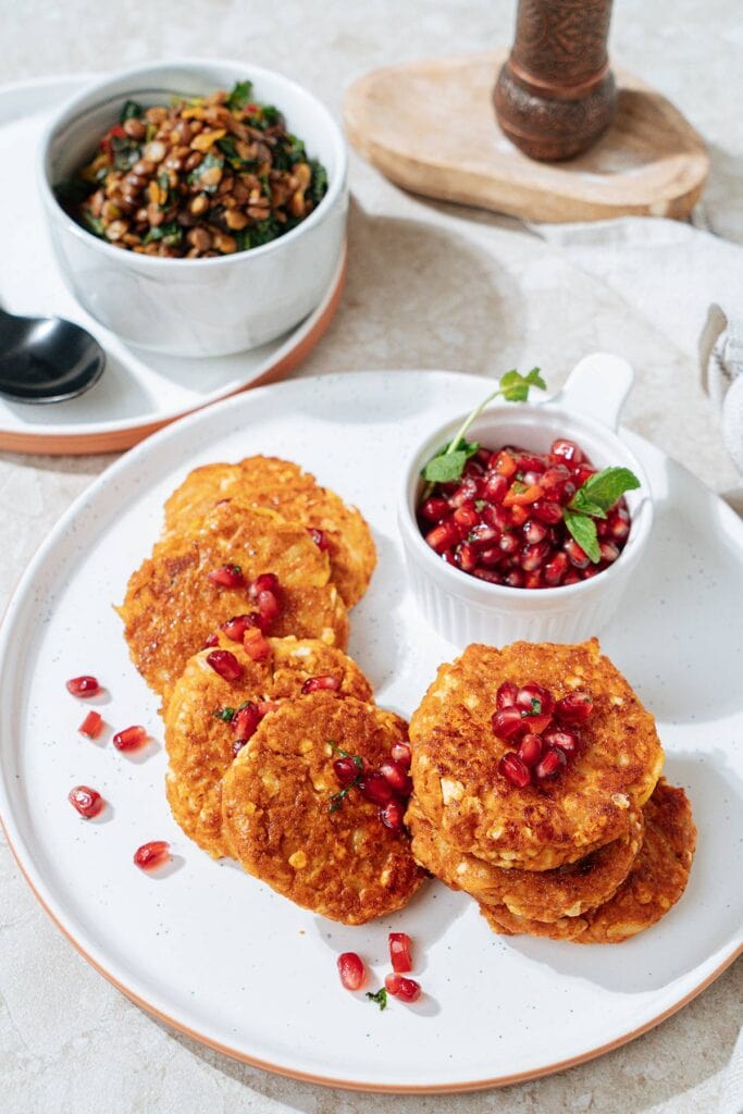 Roasted cottage cheese fritters are served on the white plate with a small dipping bowl of pomegranate salsa near them. Next to it there is a lentil chard side is in the small white bowl | Hurry The Food Up