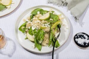 A plate of couscous and pear salad with goats cheese, on a white surface with a black fork & salt dish | Hurry The Food Up