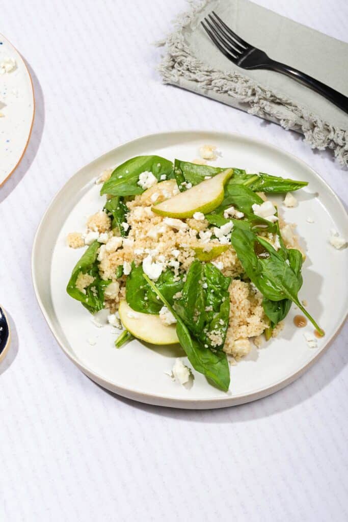 A white plate of couscous and pear salad with goats cheese, with a black fork on a napkin behind it | Hurry The Food Up