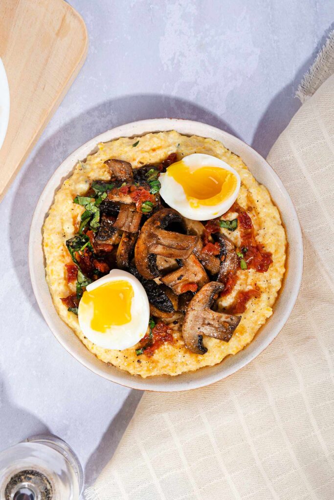 A cream bowl of polenta with mushrooms tomatoes and basil with an open soft boiled egg on top sits on a marbled surface and cream cloth next to a chopping board and pepper grinder | Hurry The Food Up