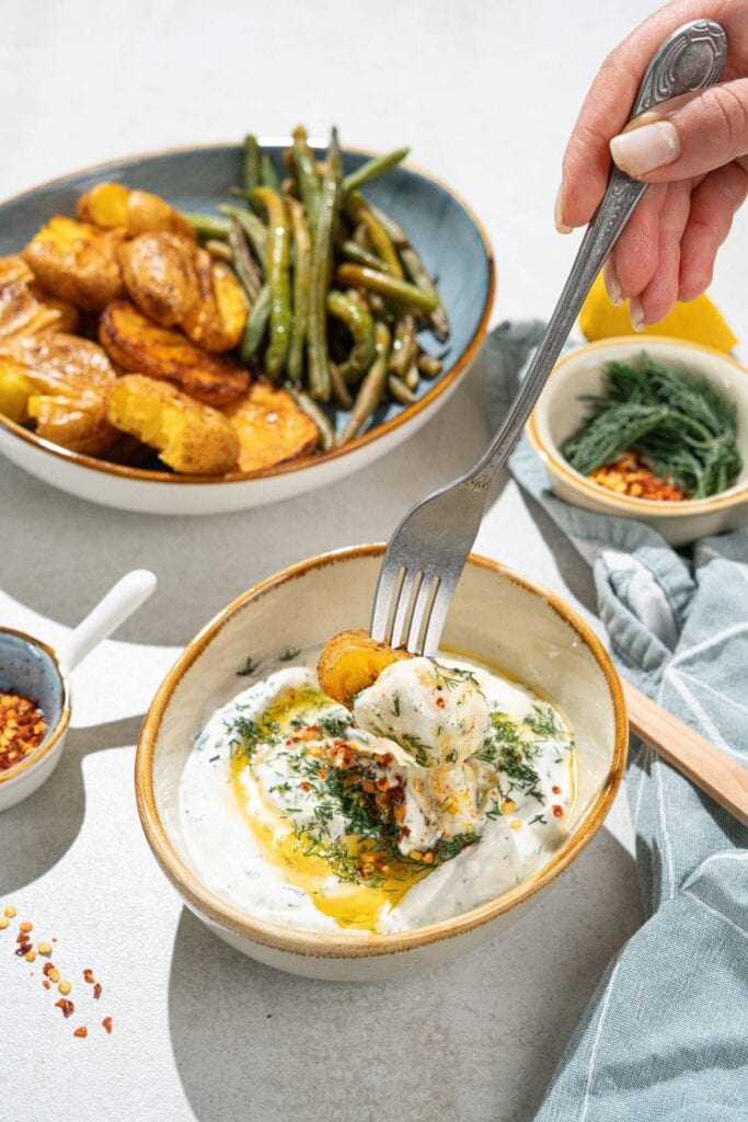 The bowl with smashed potatoes and green beans is on the grey surface. Near it, there are smaller bowls with feta dipping sauce, sweet paprika flakes, dill etc | Hurry The Food Up