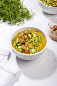 A bowl of dal khichdi on a white surface. Surrounding it are herbs, a glass bowl of salad, another bowl of khichdi and a white and brown serviette. | Hurry The Food Up