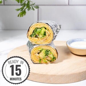 Two breakfast egg wraps in foil are on a cutting board, there is a small bowl of sour cream near them | Hurry The Food Up