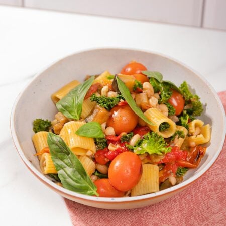 Saucy eggplant tomato pasta is in the white plate on the pink napkin that is on the table. Next to it there is a plate with basil | Hurry The Food Up