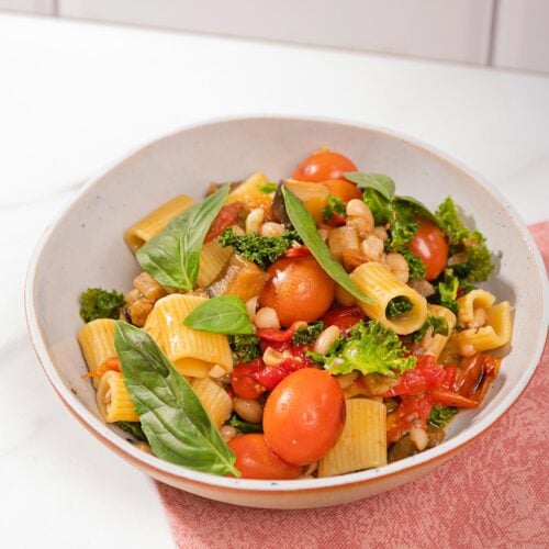 Saucy eggplant tomato pasta is in the white plate on the pink napkin that is on the table. Next to it there is a plate with basil | Hurry The Food Up