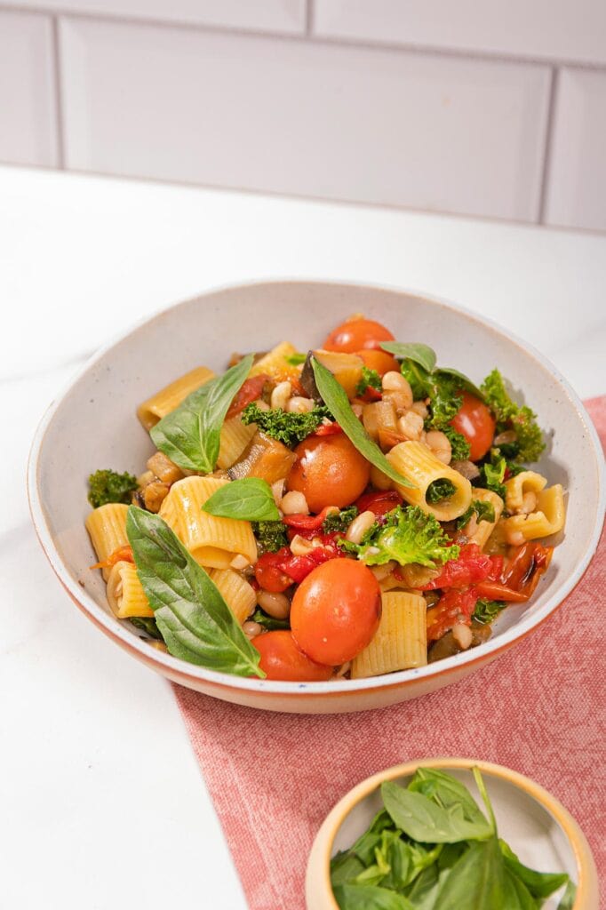 Saucy eggplant tomato pasta is in the white plate on the pink napkin that is on the table. Next to it there is a plate with basil | Hurry The Food Up