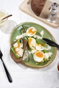 A plate of asparagus, jammy eggs, sour cream sauce and dark bread in the process of being eaten for breakfast| Hurry The Food Up