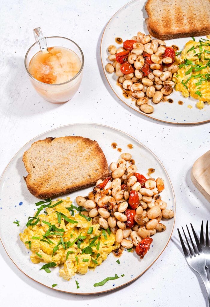 The breakfast is served with whole grain toasts on two plates that are on the white table with two forks and a cup of coffee with milk | Hurry The Food Up