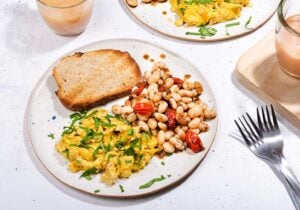 The breakfast is served with whole grain toast on a plate that is on the white table with two forks and a cup of coffee with milk and tea | Hurry The Food Up