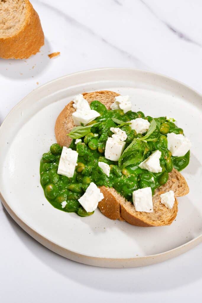 Toasted bread slice that is on the white plate is topped with beans, green sauce and crumbled cheese. There is a bread slice along it | Hurry The Food Up