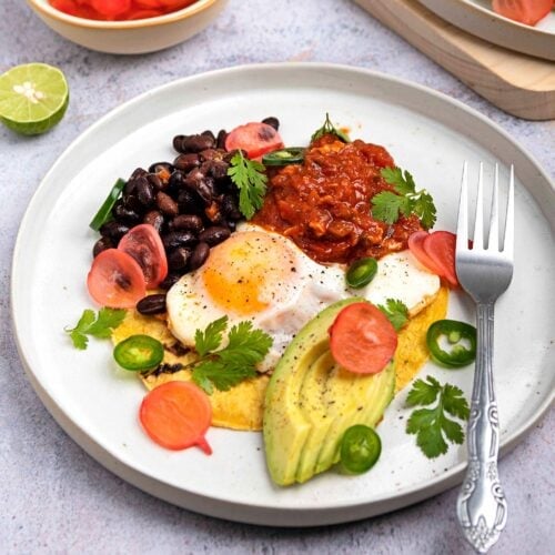 A plate of huevos rancheros on a grey surface, accompanied by a bowl of radish pickle and half a lime, with another serving of the dish in the corner of the frame | Hurry The Food Up