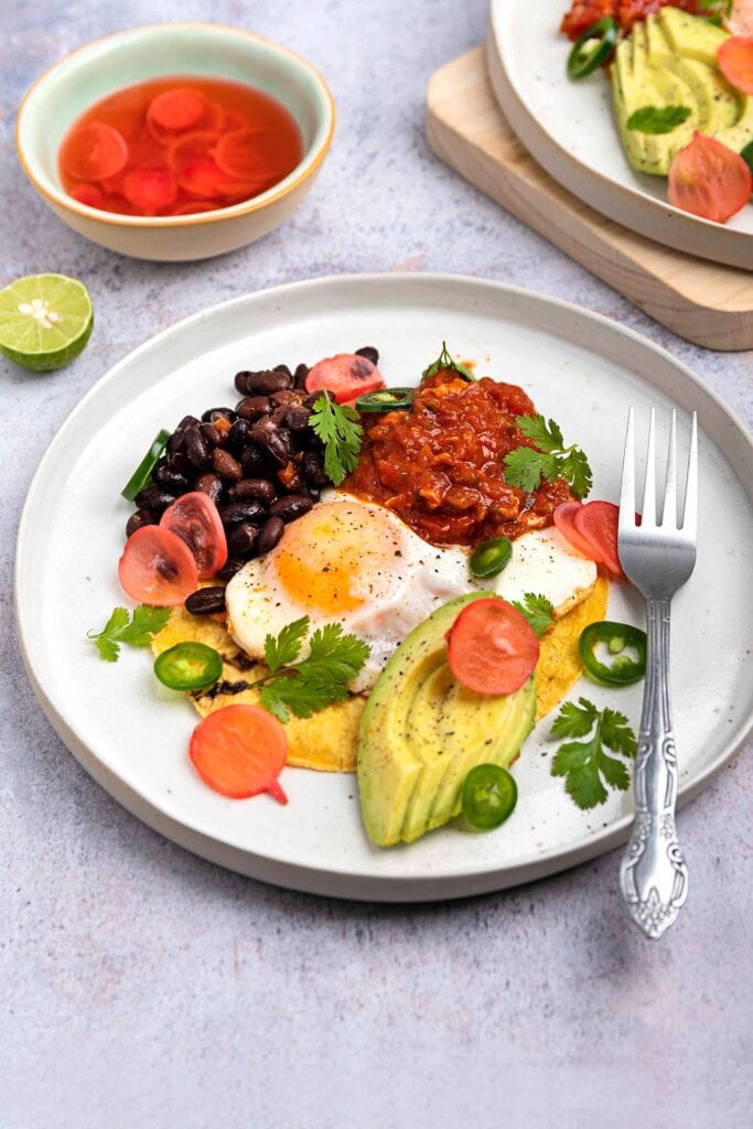 A plate of huevos rancheros on a grey surface, accompanied by a bowl of radish pickle and half a lime, with another serving of the dish in the corner of the frame | Hurry The Food Up