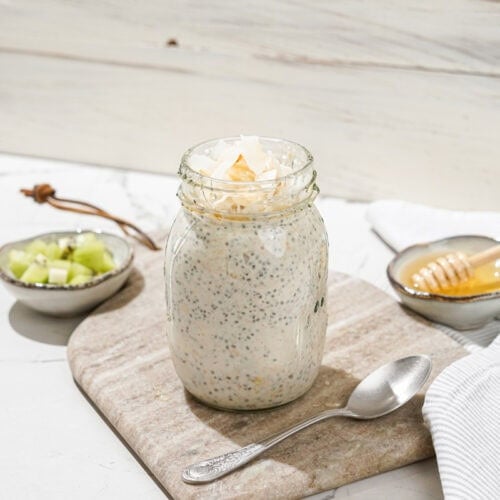 The jar with high-protein overnight oats and the spoon are on the marble сut board. Near it, there are two small bowls with kiwi and honey | Hurry The Food Up