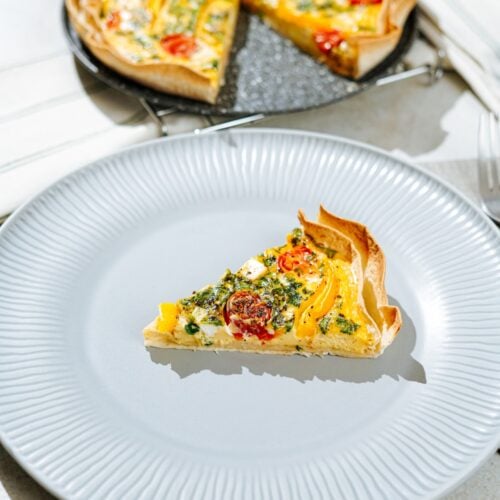 The piece of high-protein tortilla quiche lies on the white plate, and there is a fork on the right side of it. Behind the white plate, there is tortilla quiche on the black plate | Hurry The Food Up