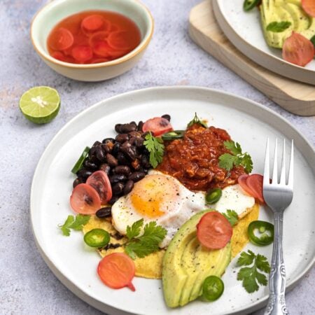 A plate of huevos rancheros on a grey surface, accompanied by a bowl of radish pickle and half a lime, with another serving of the dish in the corner of the frame | Hurry The Food Up