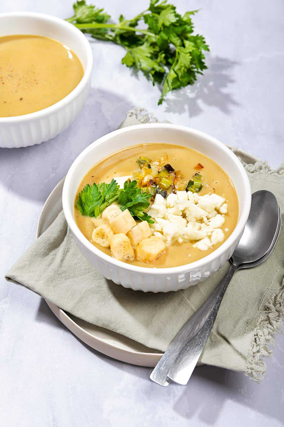 A photograph of leek and potato soup served in a white bowl, with a bunch of fresh parsley and a second bowl of soup in the top left hand corner of the image. | Hurry The Food Up