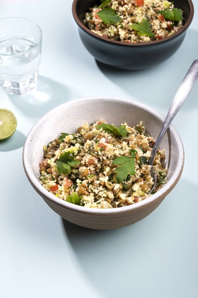 A bowl of lentil tabbouleh with a spoon on a plain surface. Visible at the top of the image is another bowl of tabbouleh , and between the two to the left is a glass of water and a slice of lime | Hurry The Food Up