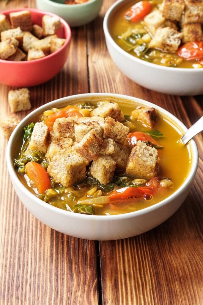 Two bowls of lentil and vegetable soup are on a wooden table. Behind them is a bowl of croutons and a smaller bowl of spices. Behind them is a sprig of rosemary. In the foreground a spoonful of lentil vegetable soup is held up | Hurry The Food Up