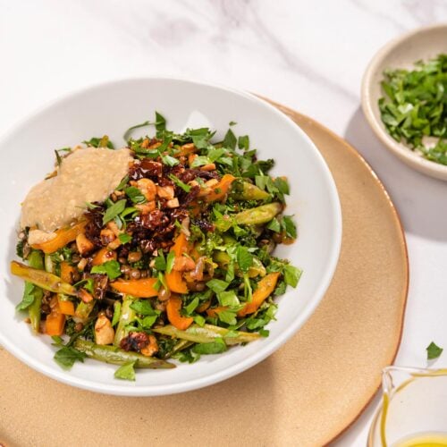 Lentils with Vegetables in Tahini Sauce are served in the white plate that stands on the beige plate that is on the table. Next to it there is a plate with chopped parsley and olive oil | Hurry The Food Up