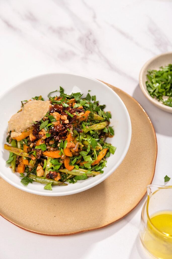 Lentils with Vegetables in Tahini Sauce are served in the white plate that stands on the beige plate that is on the table. Next to it there is a plate with chopped parsley and olive oil | Hurry The Food Up