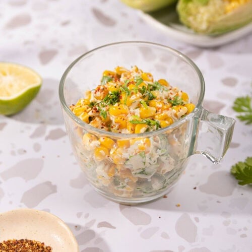 Mexican corn salad is in the glass cup on the table. Next to it there are a white plate with two corn cobs, lime half, cilantro leaves, and a plate with red pepper flakes | Hurry The Food Up