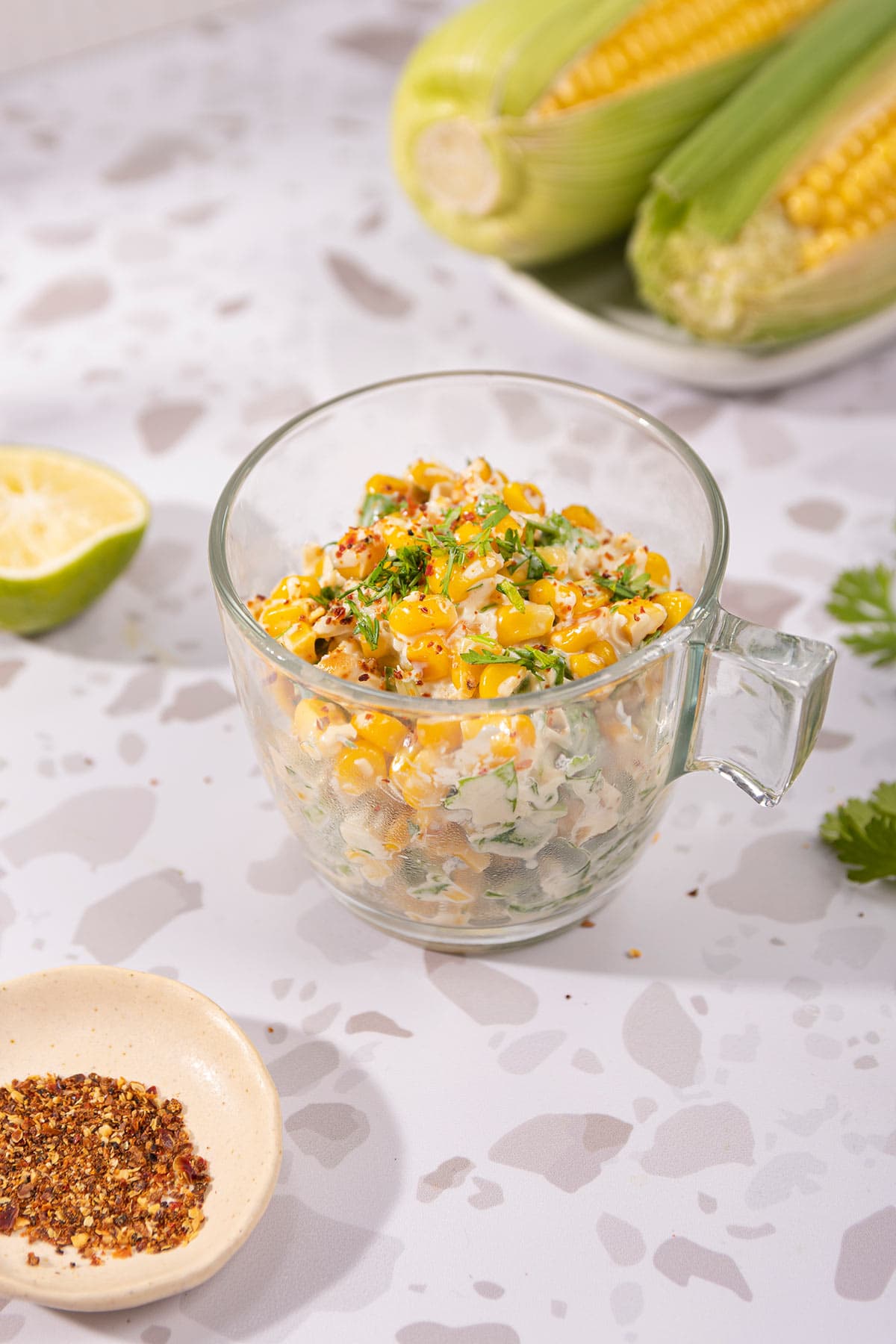 Mexican corn salad is in the glass cup on the table. Next to it there are a white plate with two corn cobs, lime half, cilantro leaves, and a plate with red pepper flakes | Hurry The Food Up