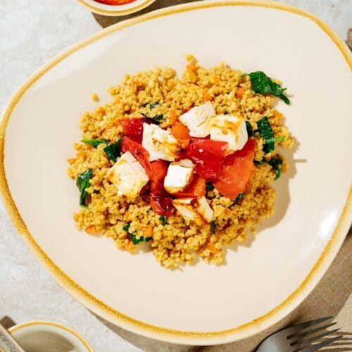 Easy high-protein millet pilaf is on the light plate with baked feta on th top of it. Near it there are the dish with baked vegetablea and feta, two spoons, fork etc | Hurry The Food Up