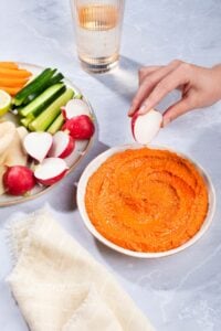 Someone is dipping a half of radish into the bowl with muhammara dip. There are a plate with vegetables and flatbreads, a glass of water and a tea towel on a side | Hurry The Food Up