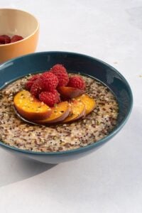 Fruity quinoa porridge is in the bowl served with pieces of peach and raspberries on the light surface. There is a bowl with raspberries behind it | Hurry The Food Up