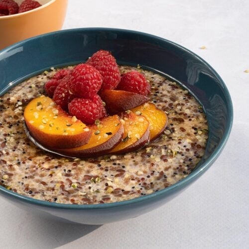 Fruity quinoa porridge is in the bowl served with pieces of peach and raspberries on the light surface. There is a bowl with raspberries behind it | Hurry The Food Up