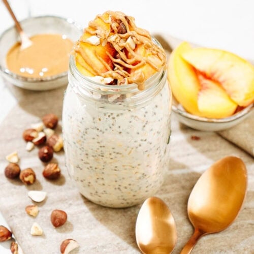 The Mason jar with overnight oats is on the marble surface. There are also some hazelnuts, two bronze spoons, a tiny bowl with peanut butter and a small dish with some slices of peach | Hurry The Food Up