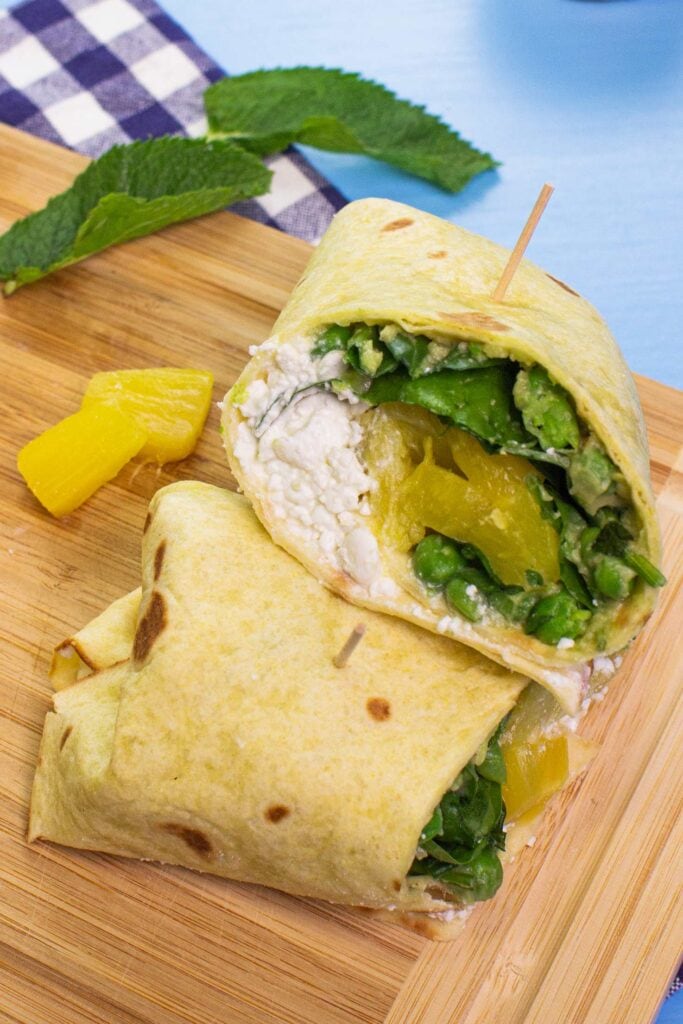 The two pieces of Pineapple Cottage Breakfast Burritos, two pieces of pineapple and two mint leaves are on the chopping board on a towel on the blue table | Hurry The Food Up