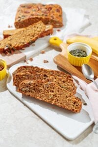 The three pieces of deeply golden brown protein banana bread lie on the white board. Around it, there are small bowls with pumpkin seeds and pecans, a spoon, a banana, and other pieces of banana bread | Hurry The Food Up