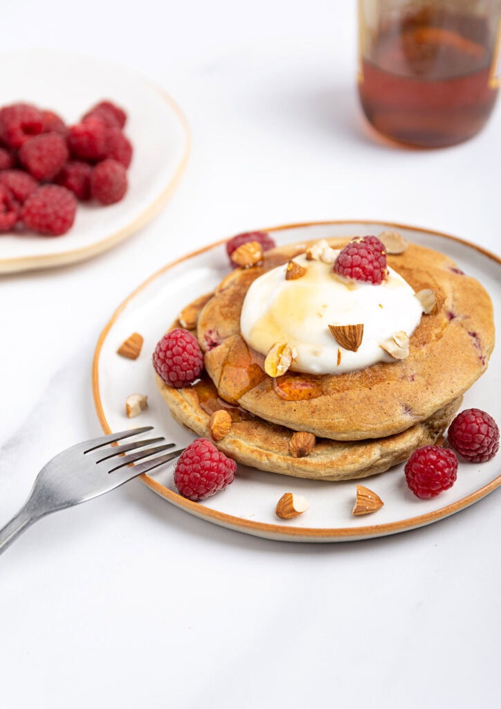 Raspberry low calorie pancakes are served on the white plate with fork and topped with yogurt, almonds and raspberries | Hurry The Food Up