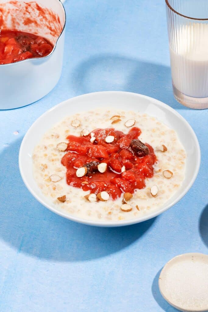 A bowl of rhubarb and bulgur porridge, with plates of sugar, chopped almonds, pot with topping and a glass of milk | Hurry The Food Up