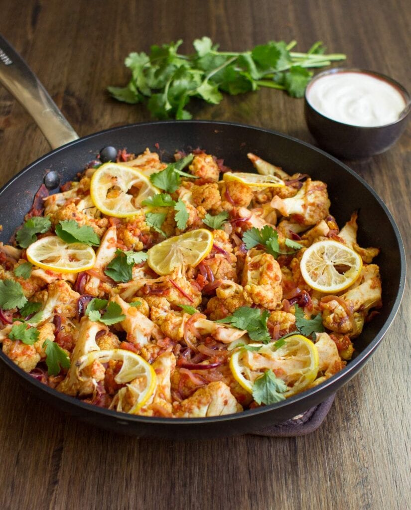 Roasted Cauliflower Curry is in the pan on the wooden surface. Next to it there is a plate with yogurt and fresh sprig cilantro | Hurry The Food Up