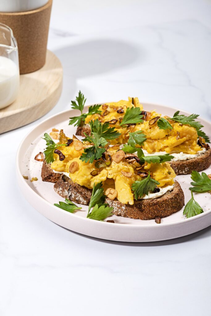 A plate of scrambled eggs taken from a side-long angle, with a milk jug and the bottom of a coffee mug visible in the lefthand corner | Hurry The Food Up