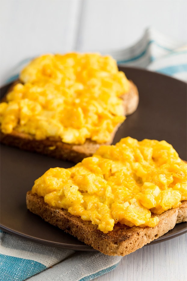 Two pieces of bread with perfect scrambled eggs with cheese lie on the black plate that is on the napkin | Hurry The Food Up