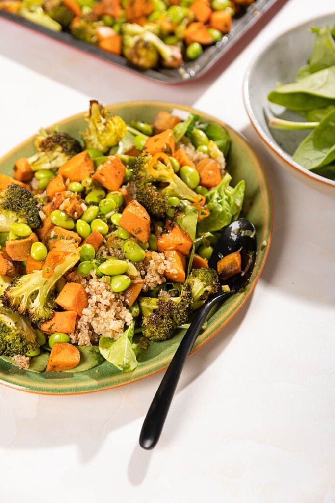 On a cream table is a sweet potato and broccoli salad on a large oval platter with a serving spoon. In the background is a black baking tray of roasted vegetables and a bowl of spinach leaves | Hurry The Food Up