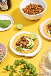 A sweet potato taco is assembled on a plate on a yellow surface. Around it are other plates and bowls, one with avocado, one with sweet potato and chickpeas, some pico de gallo, sour cream etc. | Hurry The Food Up